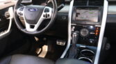 Driver’s seat view of a Ford interior: steering wheel with built‑in controls, dashboard gauges, and a center touchscreen displaying a navigation map alongside climate controls and gear selector in the foreground