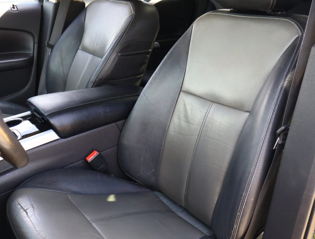 Front seats of a car with black leather upholstery and white stitching, center console visible around them.