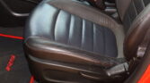 Close-up of a black leather car seat with red contrast stitching, buckled seatbelt nearby, and dark interior trim visible. Note: includes a red-accented floor mat edge near the seat.