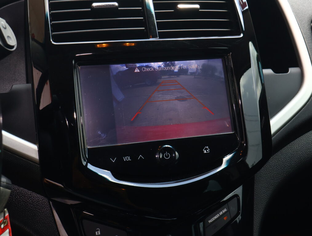 In-car infotainment screen showing a rearview camera feed with orange parking guide lines and the message 'Check Surroundings For Safety'.
