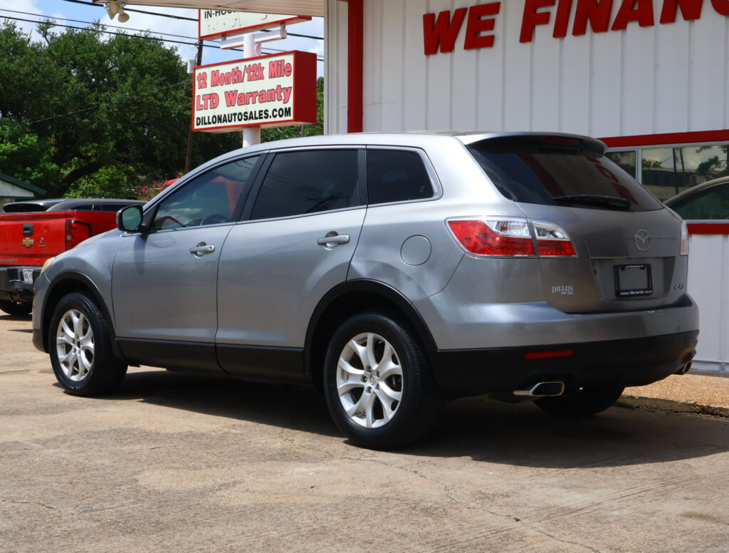 Silver SUV parked in front of a white and red car dealership building with a bold 'WE FINANCE' sign and a large roadside sign advertising 12-month/12k mile LTD warranty next to green trees.