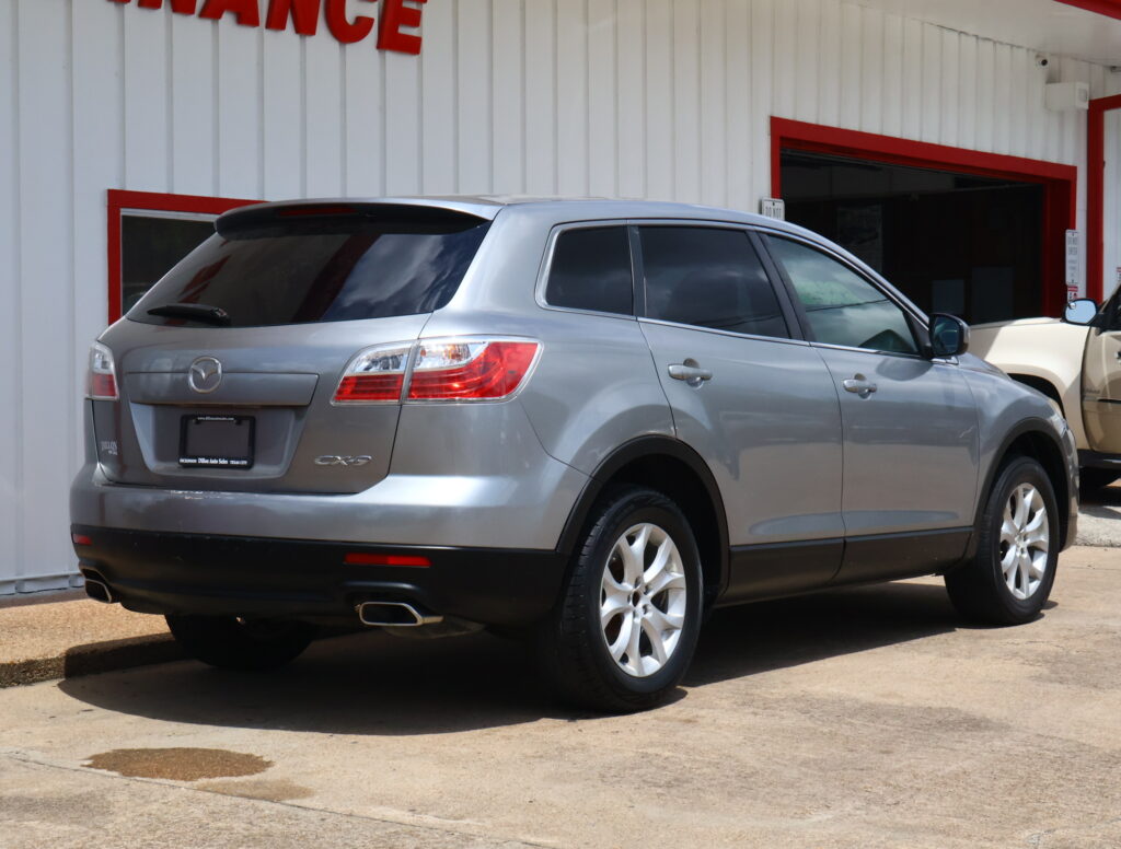 Rear three-quarters view of a silver Mazda CX-9 SUV parked in front of a white building with red trim.