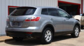 Rear three-quarters view of a silver Mazda CX-9 SUV parked in front of a white building with red trim.