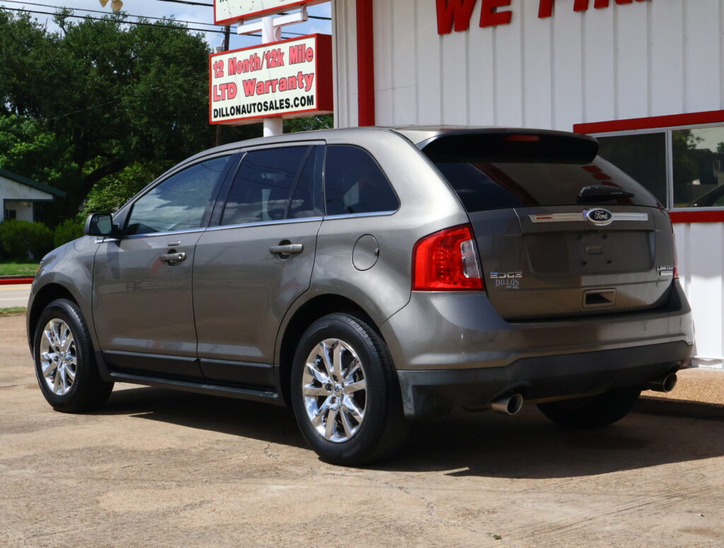 Grey Ford Edge SUV parked in front of a white and red dealership building with a warranty sign above: '12 Month/12k Mile LTD Warranty'