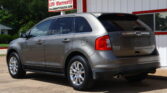 Grey Ford Edge SUV parked in front of a white and red dealership building with a warranty sign above: '12 Month/12k Mile LTD Warranty'