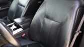 Black leather car seats with red stitching; visible wear and cracking on the driver's seat. Concise interior shot showing center console and seat belt buckle.