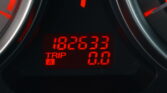 Close-up of a car dashboard with a red digital display showing trip/odometer readings; surrounding gauge needles are faintly visible in the dark.