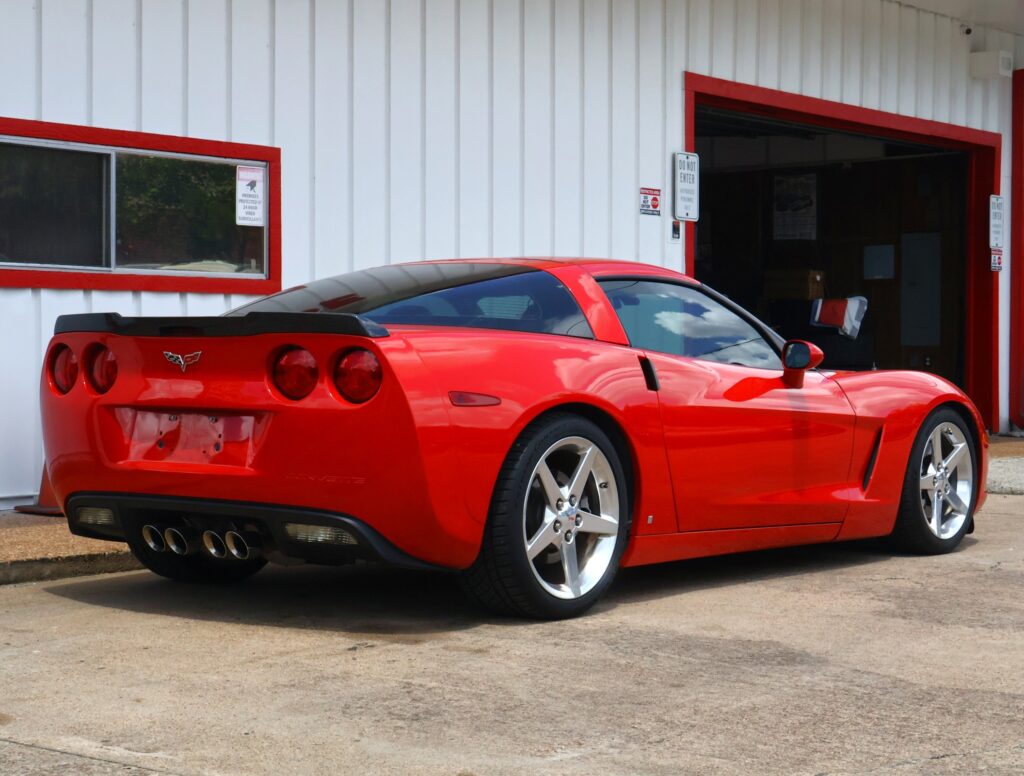 Red Chevrolet Corvette parked beside a white building with red window/door trim.