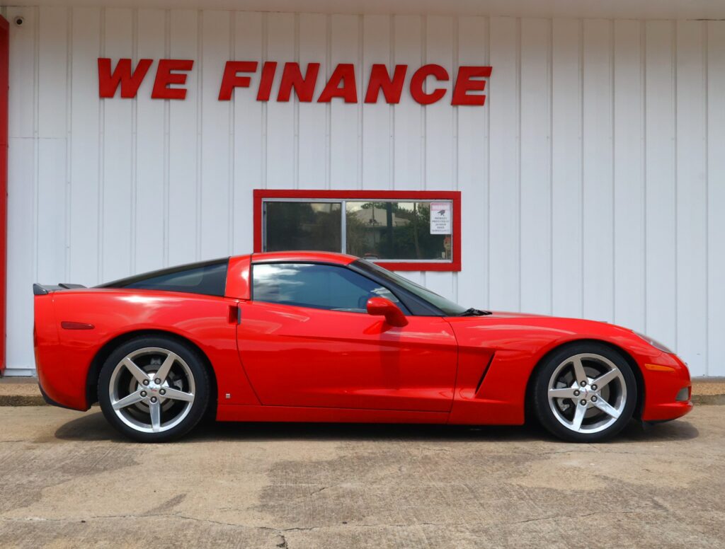 Bright red sports car parked in front of a white corrugated building with a large red 'WE FINANCE' sign above a window.