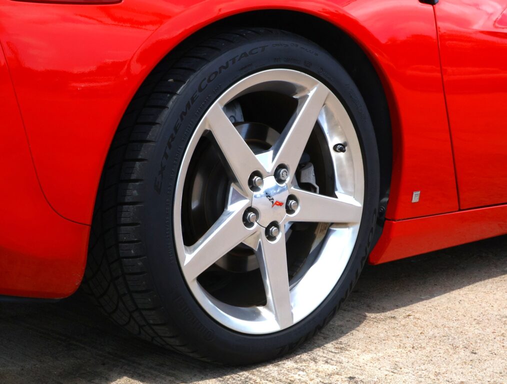 Close-up of a five-spoke silver alloy wheel on a red car, with a black tire and visible Corvette center emblem in the wheel.