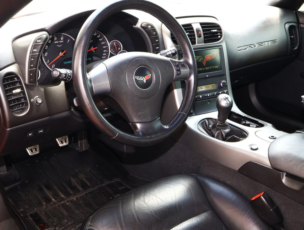 Close-up of a black leather car interior featuring a steering wheel with the Corvette emblem, dashboard gauges, and center console.