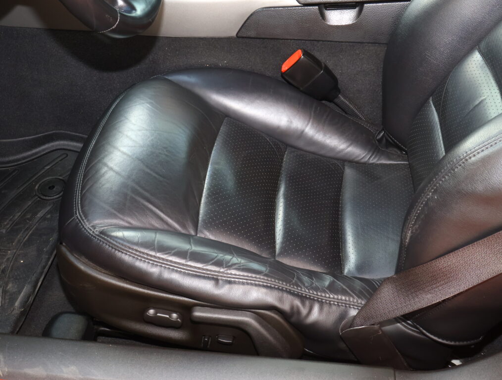 Black leather car seat with small perforations and a red seat belt buckle resting on the seat, shown in a car interior with the floor mat nearby.