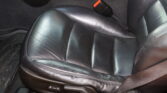 Black leather car seat with small perforations and a red seat belt buckle resting on the seat, shown in a car interior with the floor mat nearby.