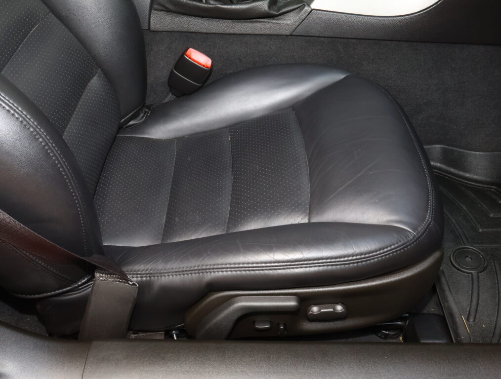 Close-up of a black leather car seat with a perforated pattern and a red seat belt buckle nearby.