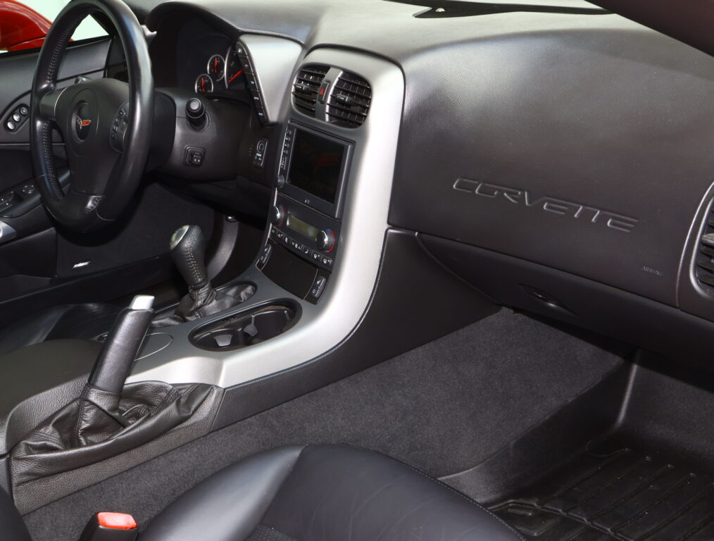 Interior view of a black Corvette cockpit with steering wheel, center console, and Corvette branding on the passenger side dash.