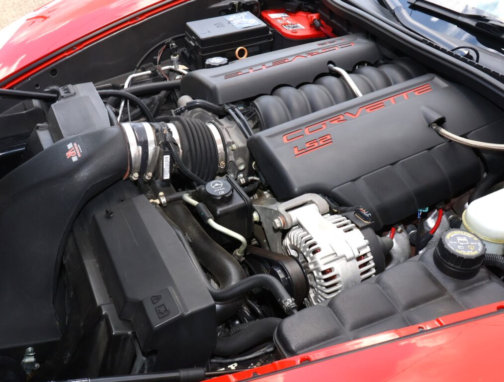 Under-hood view of a red Corvette with the LS2 V8 engine, black intake covers, and red 'CORVETTE LS2' lettering.