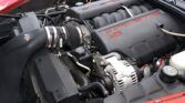 Under-hood view of a red Corvette with the LS2 V8 engine, black intake covers, and red 'CORVETTE LS2' lettering.