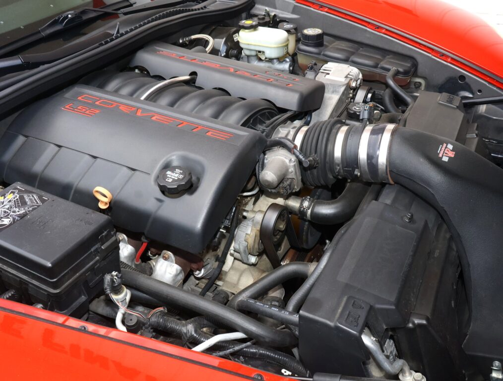 Close-up of a red Corvette engine bay, featuring the black engine covers with red 'CORVETTE' lettering and visible hoses.