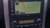 Car's navigation screen displaying a map with a red route line and a small compass overlay, surrounded by control buttons and climate dials on the dash.