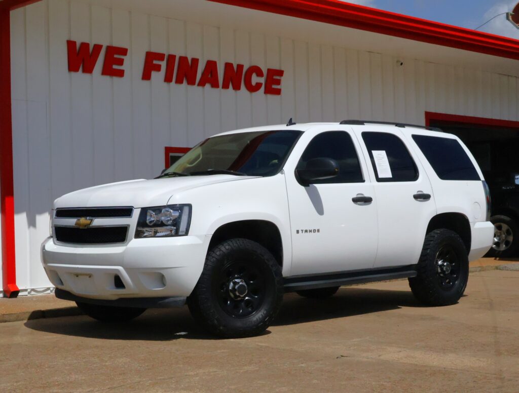 White Chevrolet Tahoe pickup/SUV parked in front of a white dealership building with a red WE FINANCE sign visible and a red trim edge.